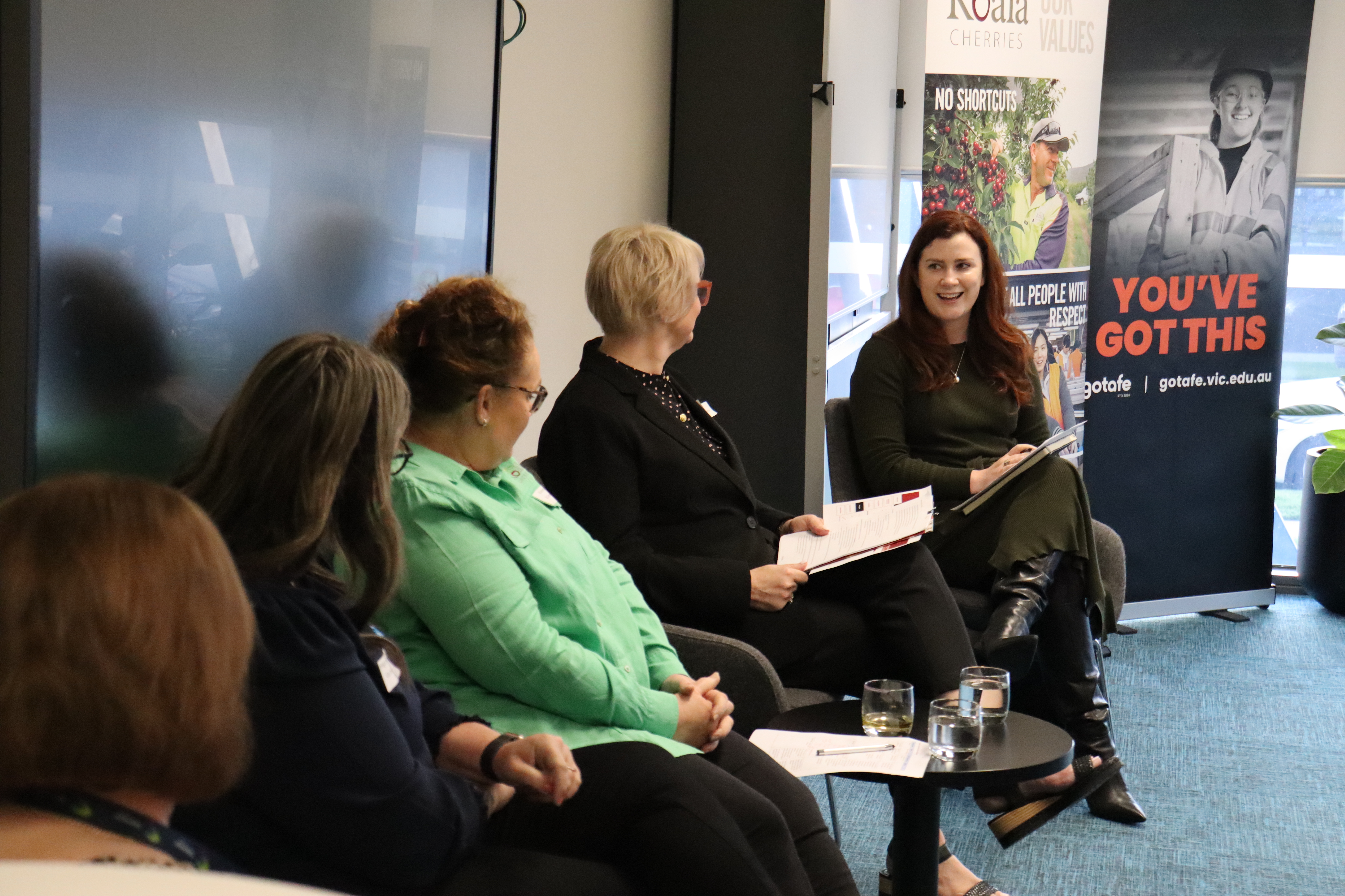 Woman with brown hair talking to four people sitting with their backs towards the camera.